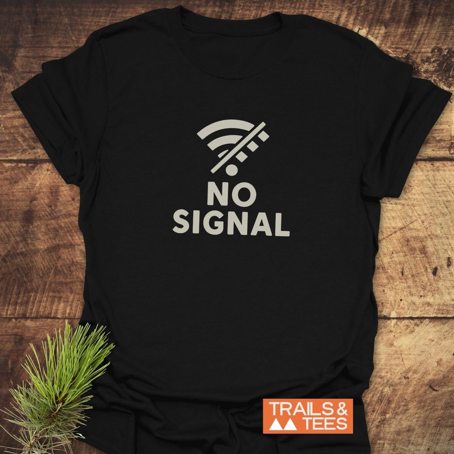 No Signal Minimalist T-Shirt featuring a crossed-out Wi-Fi icon and NO SIGNAL text, laid on wood with a pine sprig, Trails & Tees logo visible.