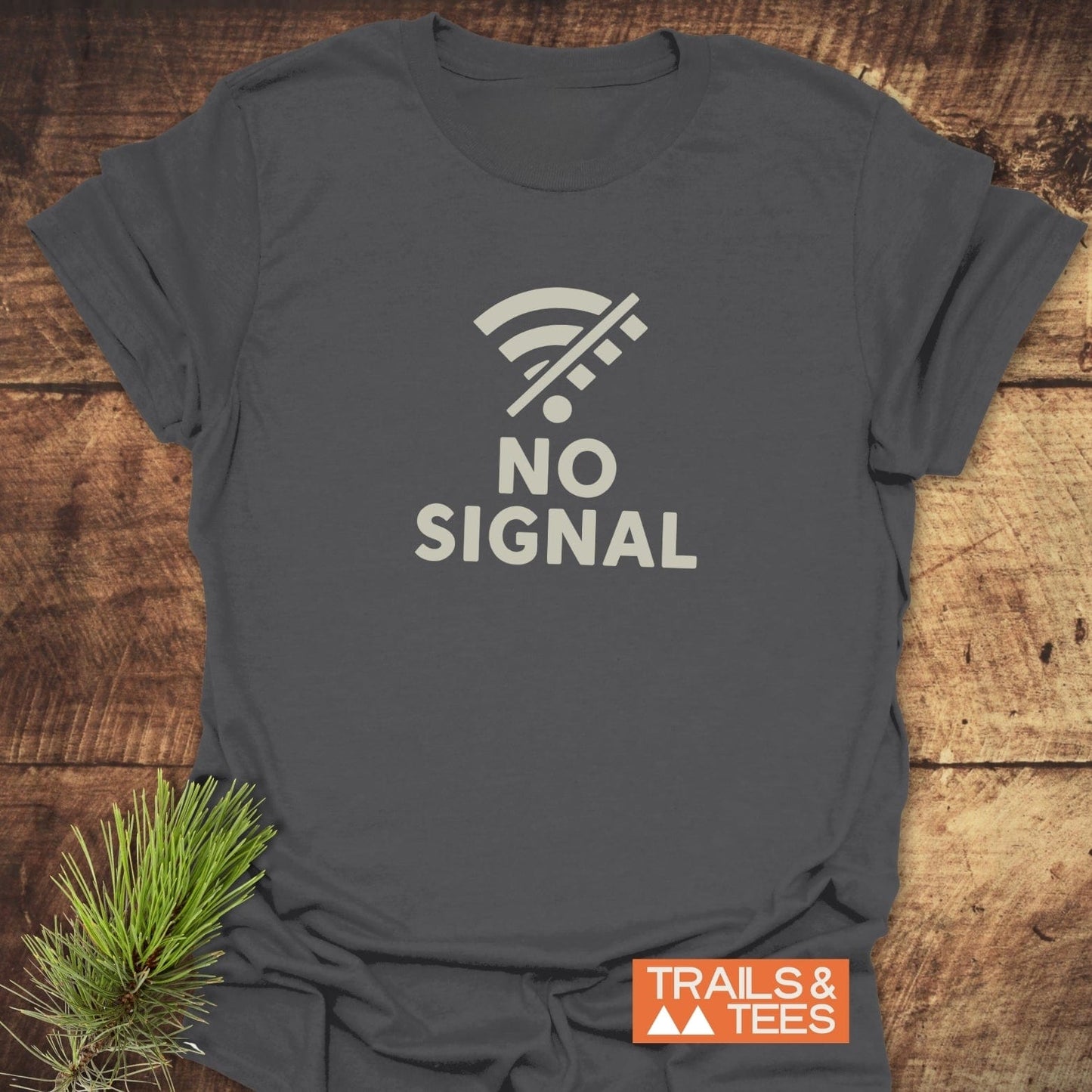 No Signal Minimalist T-Shirt on wooden surface, featuring a crossed-out Wi-Fi icon and NO SIGNAL text, next to a pine sprig and Trails & Tees logo.