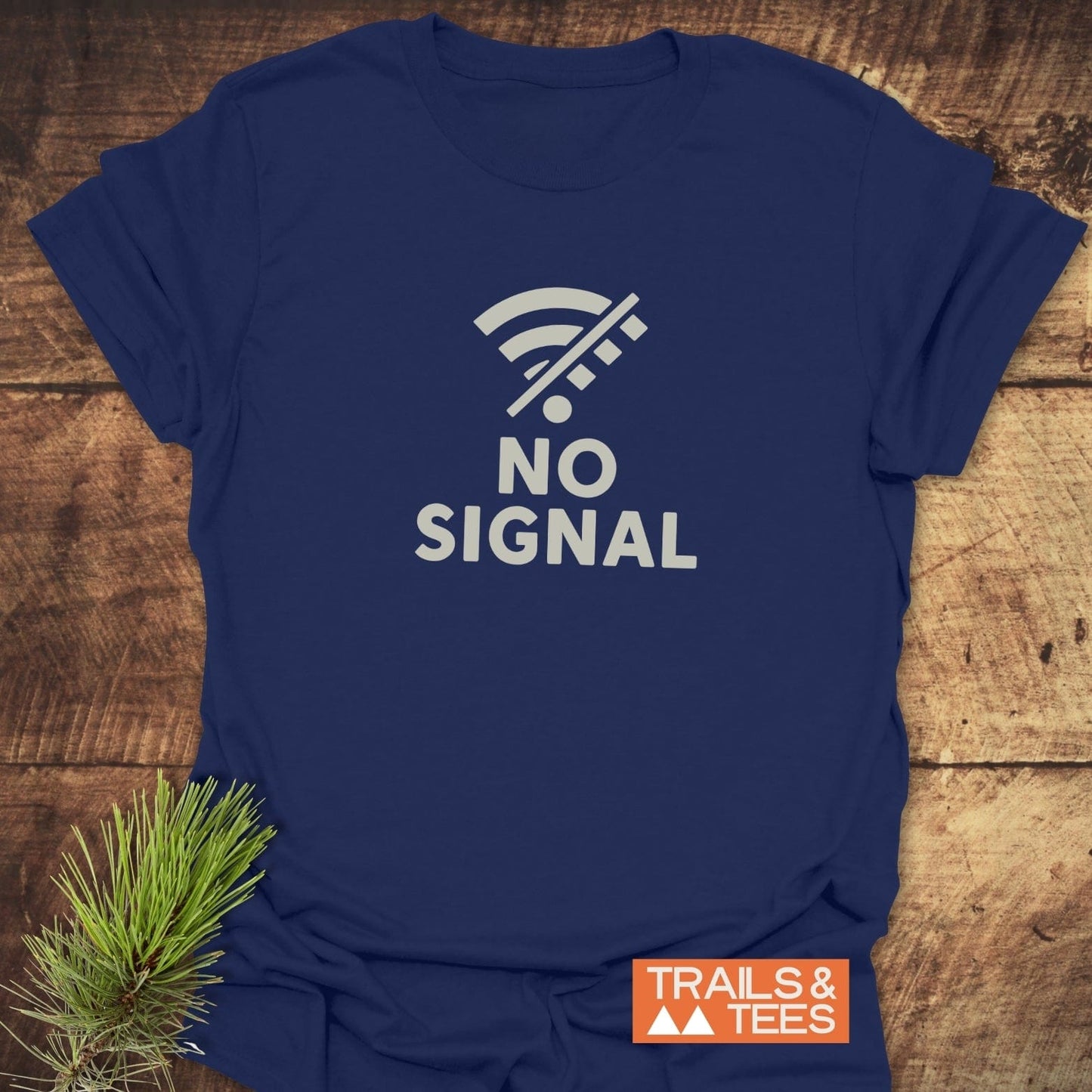 No Signal Minimalist T-Shirt featuring a crossed-out Wi-Fi icon and NO SIGNAL text, laid on wood with a pine branch, Trails & Tees logo visible.