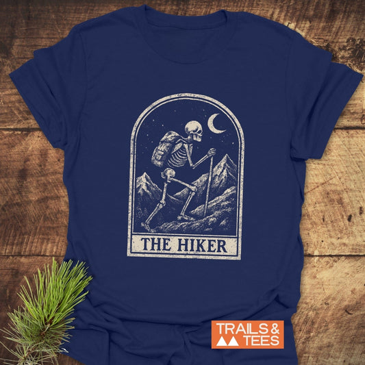 Navy-blue tee featuring distressed skeleton graphic with backpack and hiking staff, labeled THE HIKER. Includes TRAILS & TEES logo.