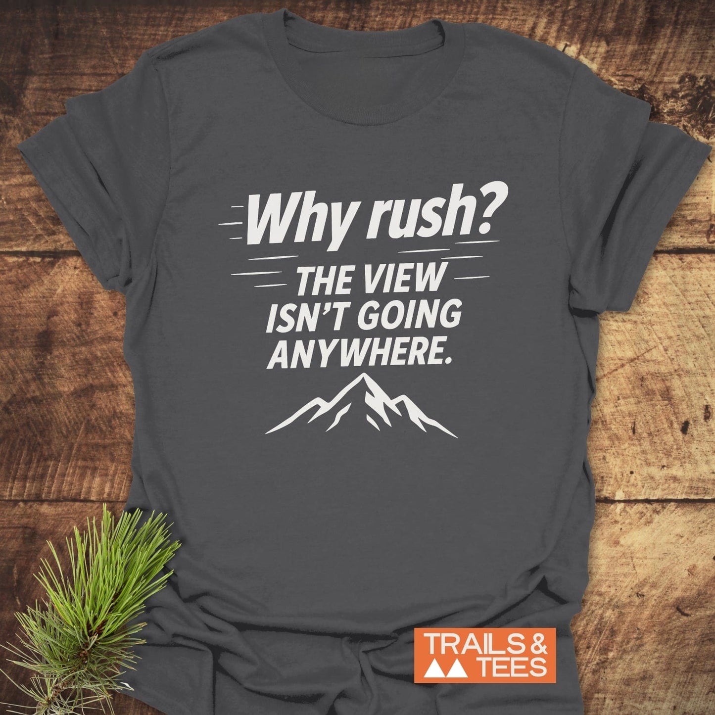 Why Rush? Slow Hikers T-Shirt on wooden surface, featuring mountain silhouette. Soft, durable cotton, pre-shrunk, no side seams for comfort. Trails & Tees logo visible.