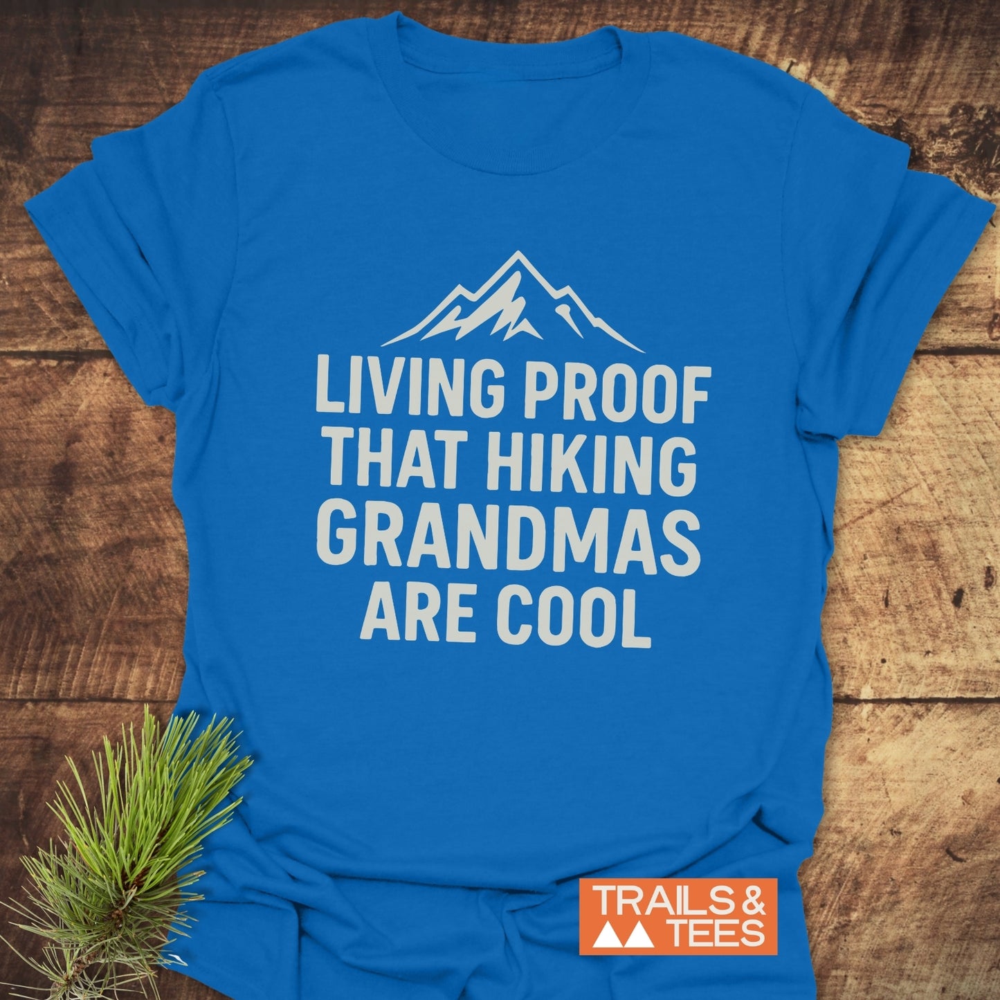 Living Proof Grandma T-Shirt featuring a mountain graphic and slogan, laid on wood. Includes Trails & Tees logo and pine sprig for an outdoor vibe.