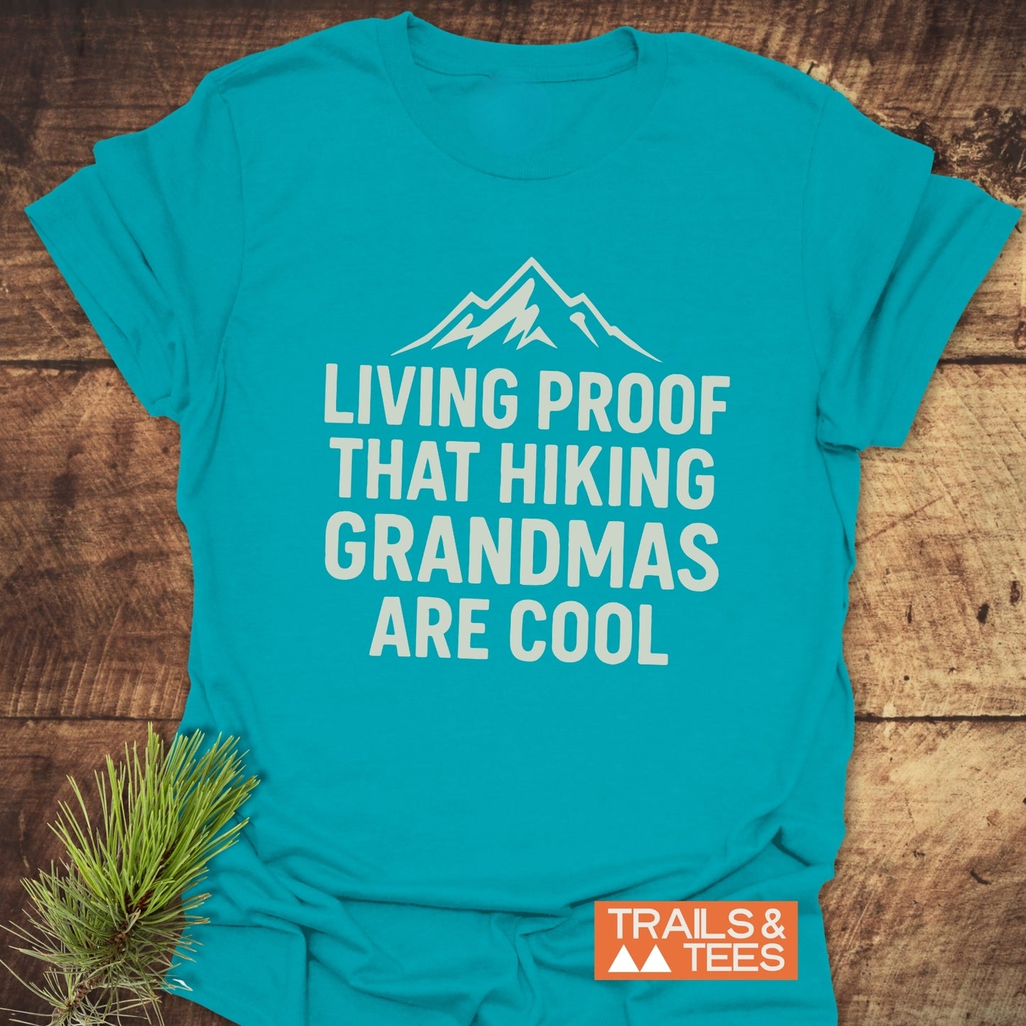 Living Proof Grandma T-Shirt featuring a white mountain graphic and slogan, laid on wood with a pine sprig, showcasing outdoor-themed comfort and durability.