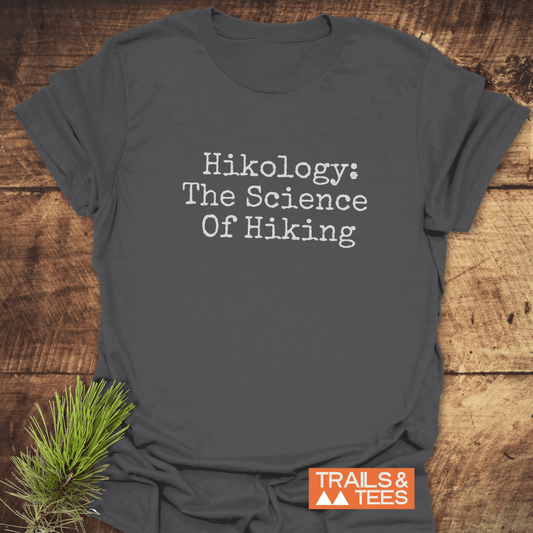 Hikology T-Shirt on wood surface, featuring The Science Of Hiking print. Includes Trails & Tees logo and pine branch accentuating outdoor theme.