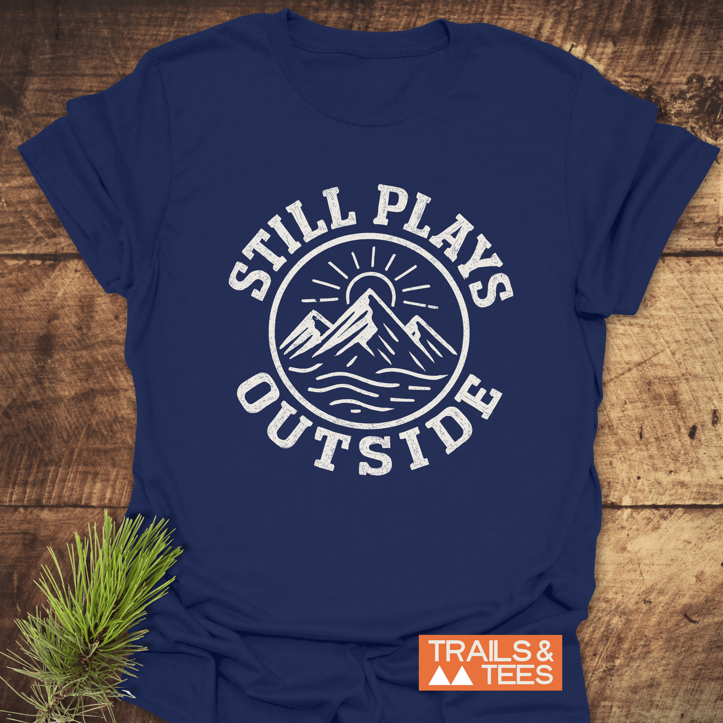 Still Plays Outside T-Shirt with a mountain and sun graphic, crafted from soft cotton, ideal for outdoor enthusiasts, lies on a wooden surface with pine accents.