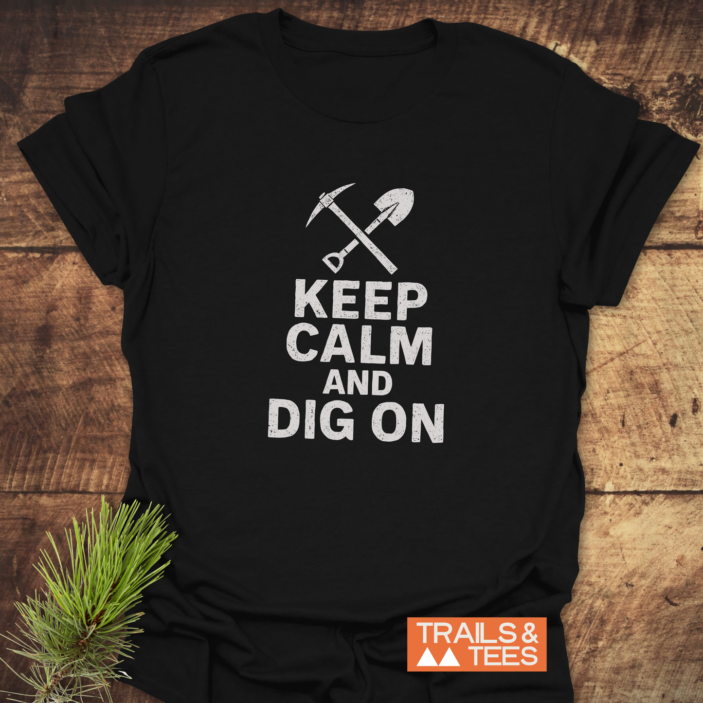 Keep Calm Rock Lover T-Shirt with crossed pickaxe and shovel graphic, slogan KEEP CALM AND DIG ON, and Trails & Tees logo, laid on wood.