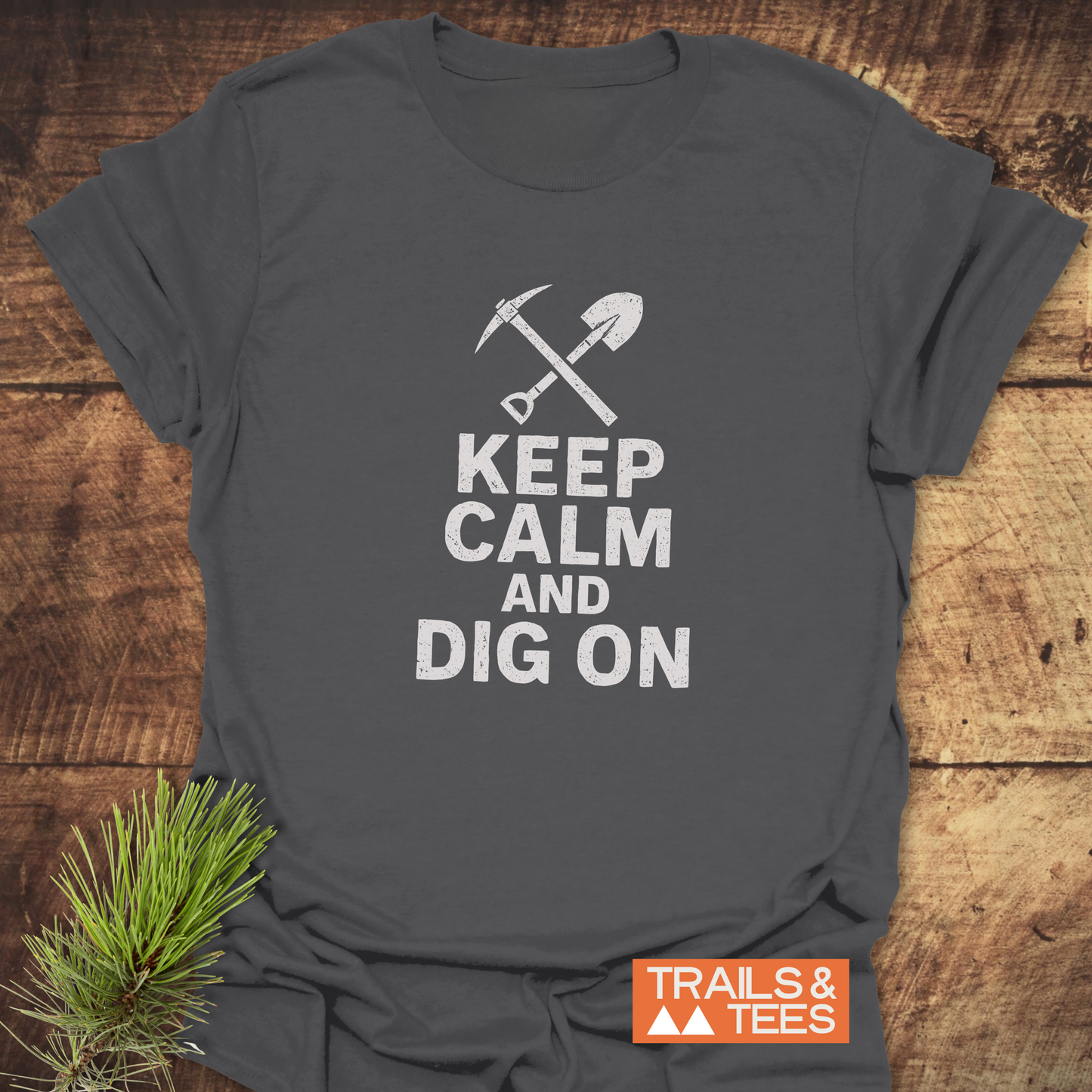 Keep Calm Rock Lover T-Shirt featuring a white pickaxe and shovel design, laid on wood. Includes TRAILS & TEES logo and pine sprig accent.