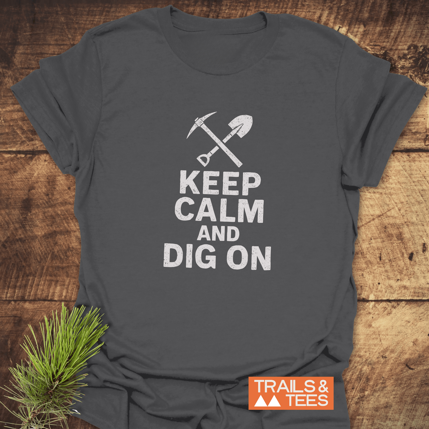 Keep Calm Rock Lover T-Shirt featuring a white pickaxe and shovel design, laid on wood. Includes TRAILS & TEES logo and pine sprig accent.