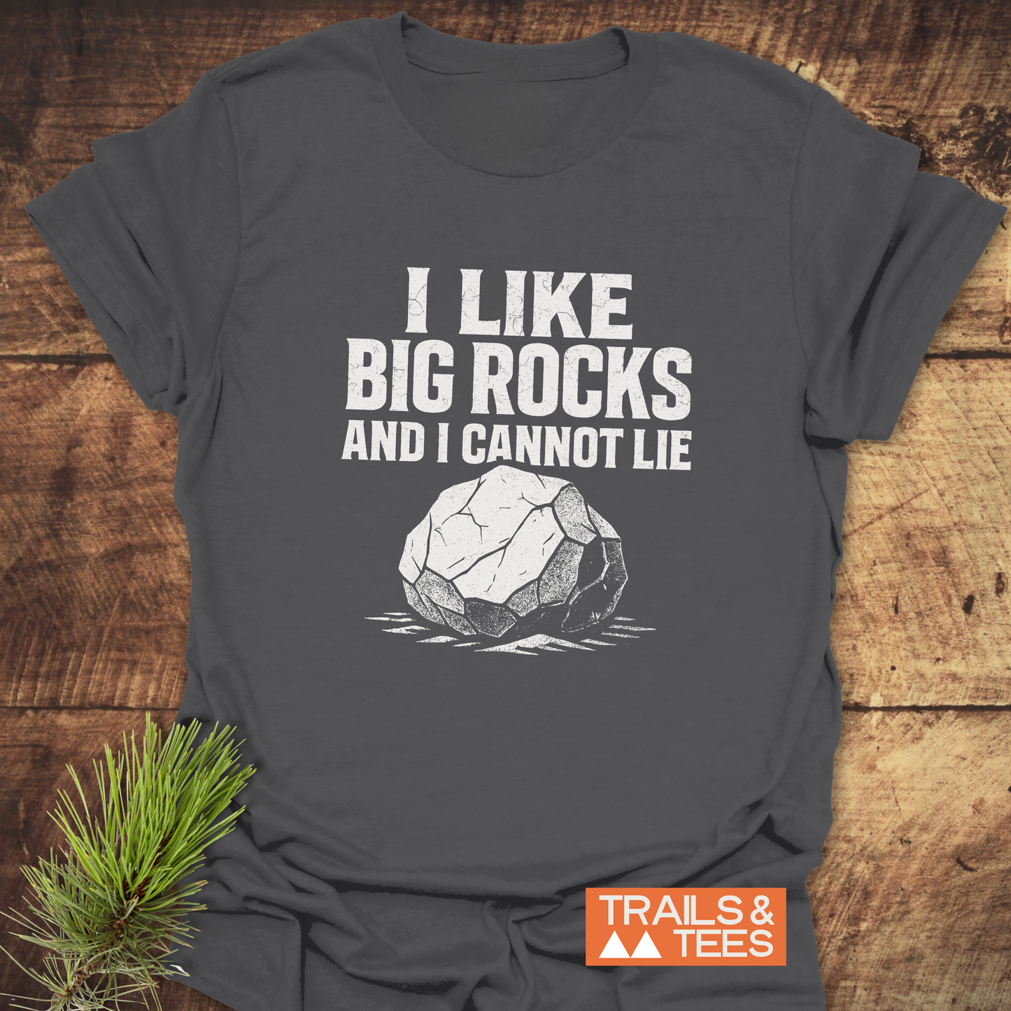 Dark-gray I Like Big Rocks T-shirt with white rock graphic, laid on wood surface. Features soft cotton, durable print, and Trails & Tees logo.