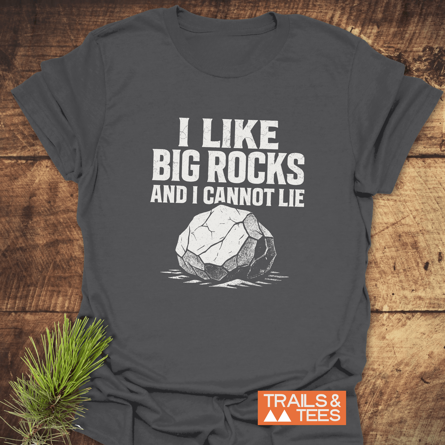 Dark-gray I Like Big Rocks T-shirt with white rock graphic, laid on wood surface. Features soft cotton, durable print, and Trails & Tees logo.