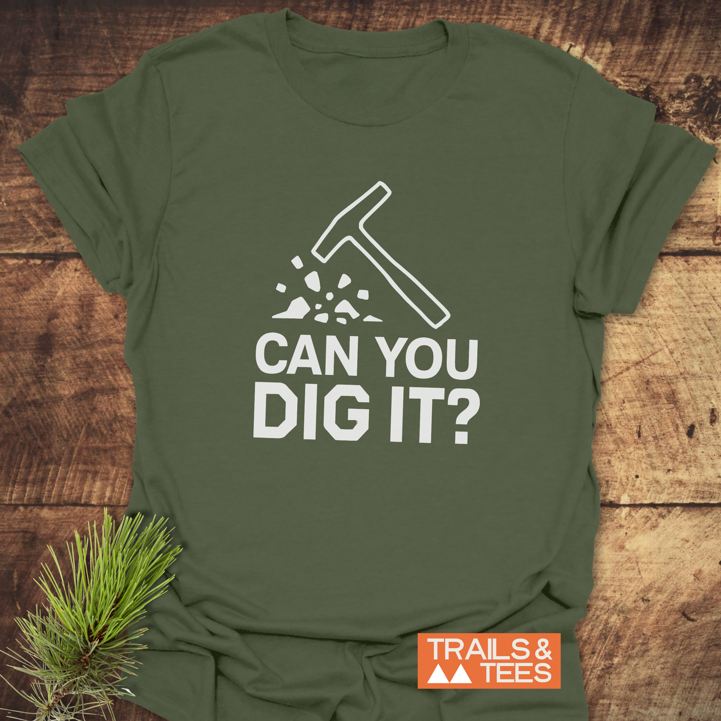 Can You Dig It? Rock Lovers T-Shirt, featuring a pick-axe graphic and slogan, displayed flat with pine decor, embodies outdoor spirit and durable comfort.