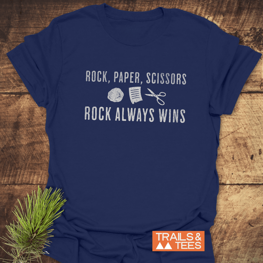 Navy-blue Rock Paper Scissors T-Shirt featuring playful graphic text and icons, showcasing durable design by Trails & Tees, laid flat on a wooden surface.