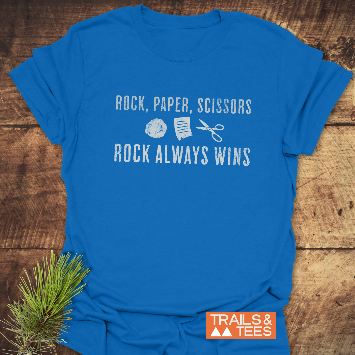 Blue Rock Paper Scissors T-Shirt with white distressed lettering and icons, featuring ROCK ALWAYS WINS slogan, orange TRAILS & TEES. logo, on wooden surface.