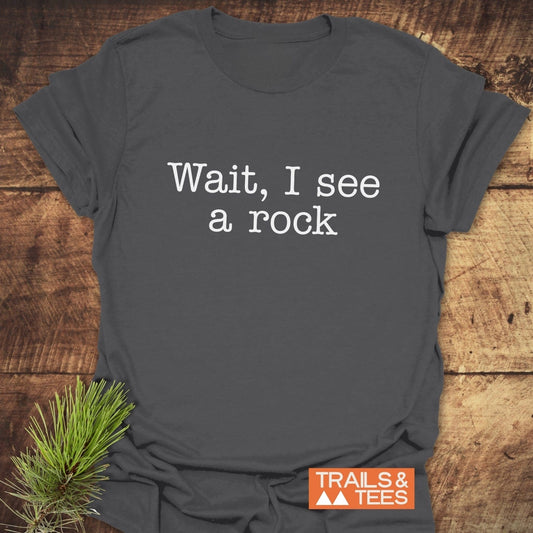 Charcoal-gray Wait, I See A Rock T-shirt with typewriter-style text, featuring TRAILS & TEES logo, styled with pine sprig; soft, durable, adventure-ready.