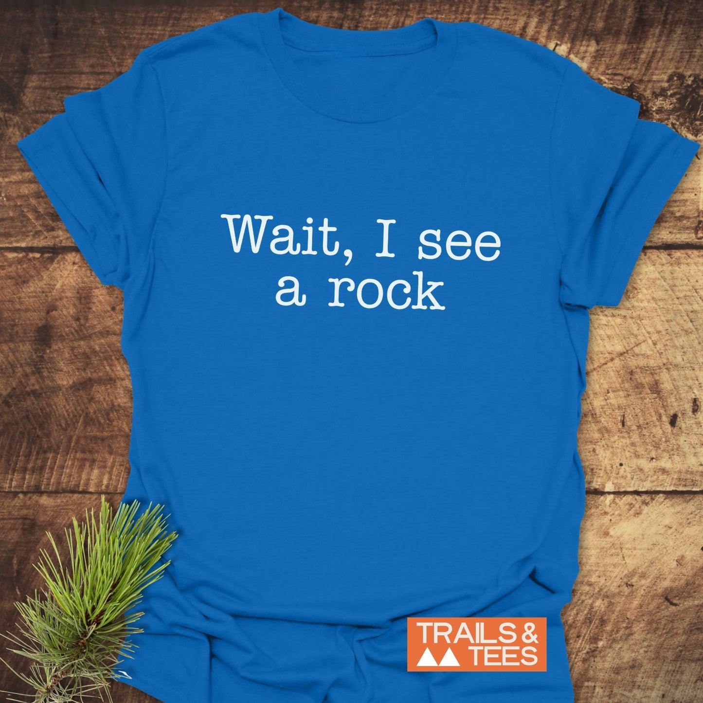 Wait, I See A Rock T-Shirt on rustic wood, featuring white typewriter-style slogan, orange TRAILS & TEES logo, and pine sprig accent for outdoor enthusiasts.
