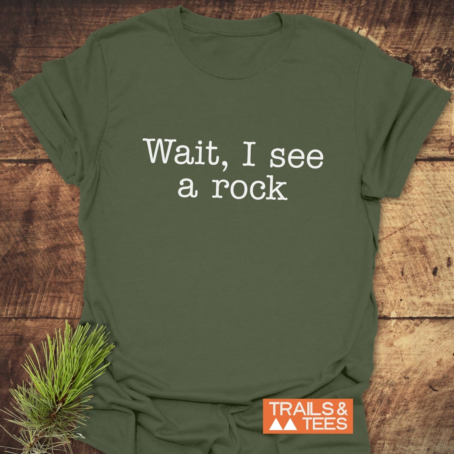 Wait, I See A Rock T-Shirt on rustic surface, featuring white text and Trails & Tees logo, styled with a pine twig for an outdoor vibe.
