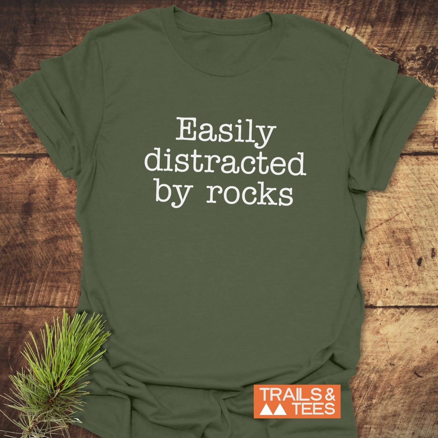 Easily Distracted Rocklover T-Shirt showing white slogan on forest-green fabric, styled with pine needles, featuring a durable design and orange TRAILS & TEES logo.