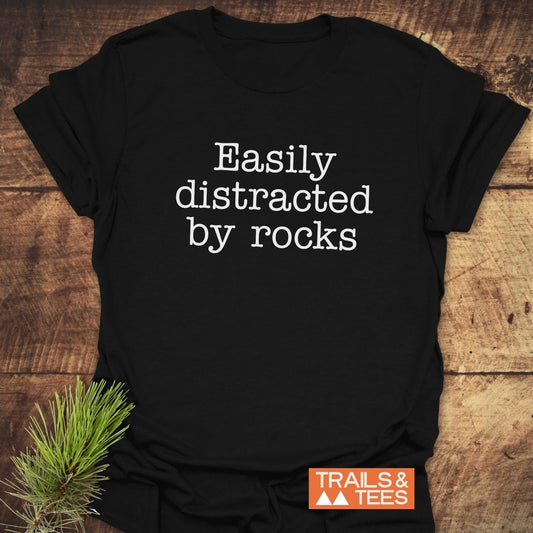 Easily Distracted Rocklover T-Shirt on wooden surface, featuring white typewriter-style slogan. Includes small “TRAILS & TEES” logo and pine sprig for styling.