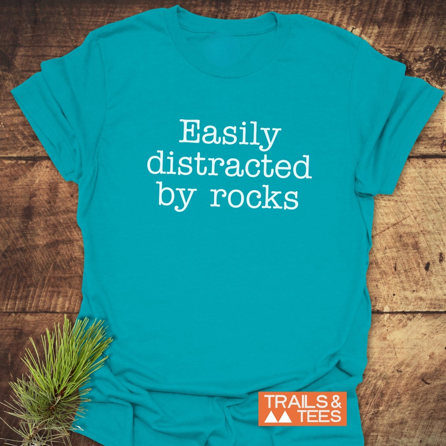 Easily Distracted Rocklover T-Shirt on wood surface, featuring white text and orange TRAILS & TEES logo, styled with a pine branch.