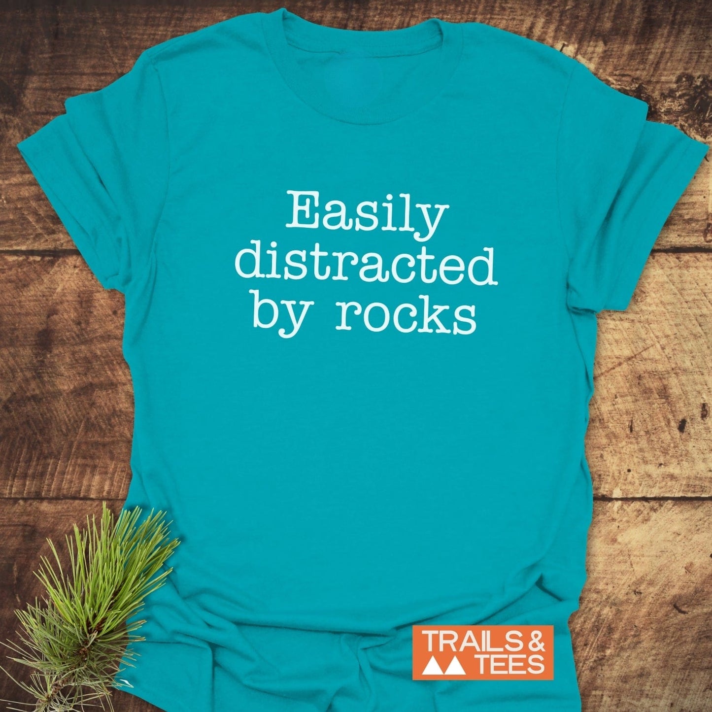 Easily Distracted Rocklover T-Shirt on wood surface, featuring white text and orange TRAILS & TEES logo, styled with a pine branch.