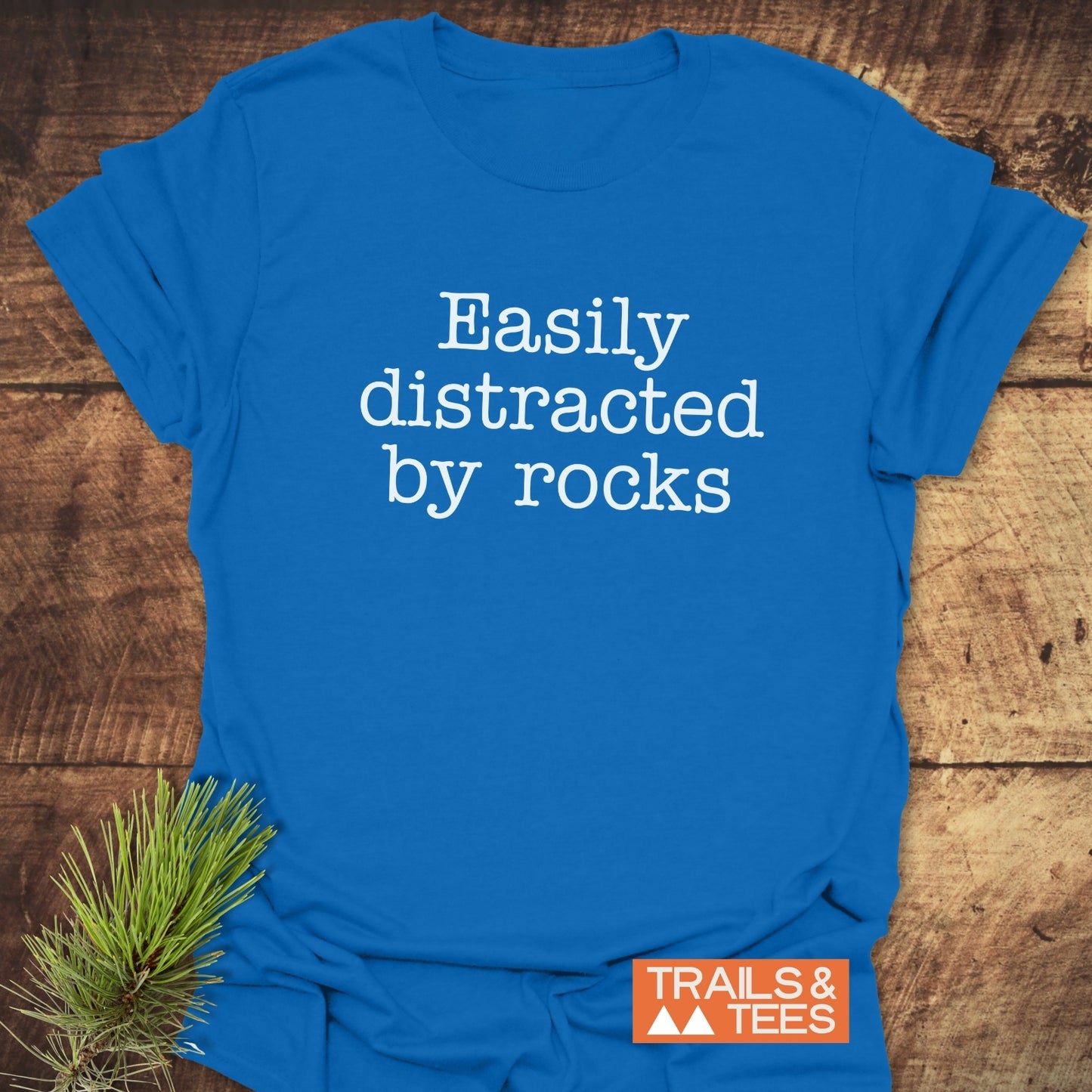 Easily Distracted Rocklover T-Shirt on rustic wood, features white typewriter-style text, pine sprig, and orange TRAILS & TEES logo, highlighting outdoor adventure spirit.