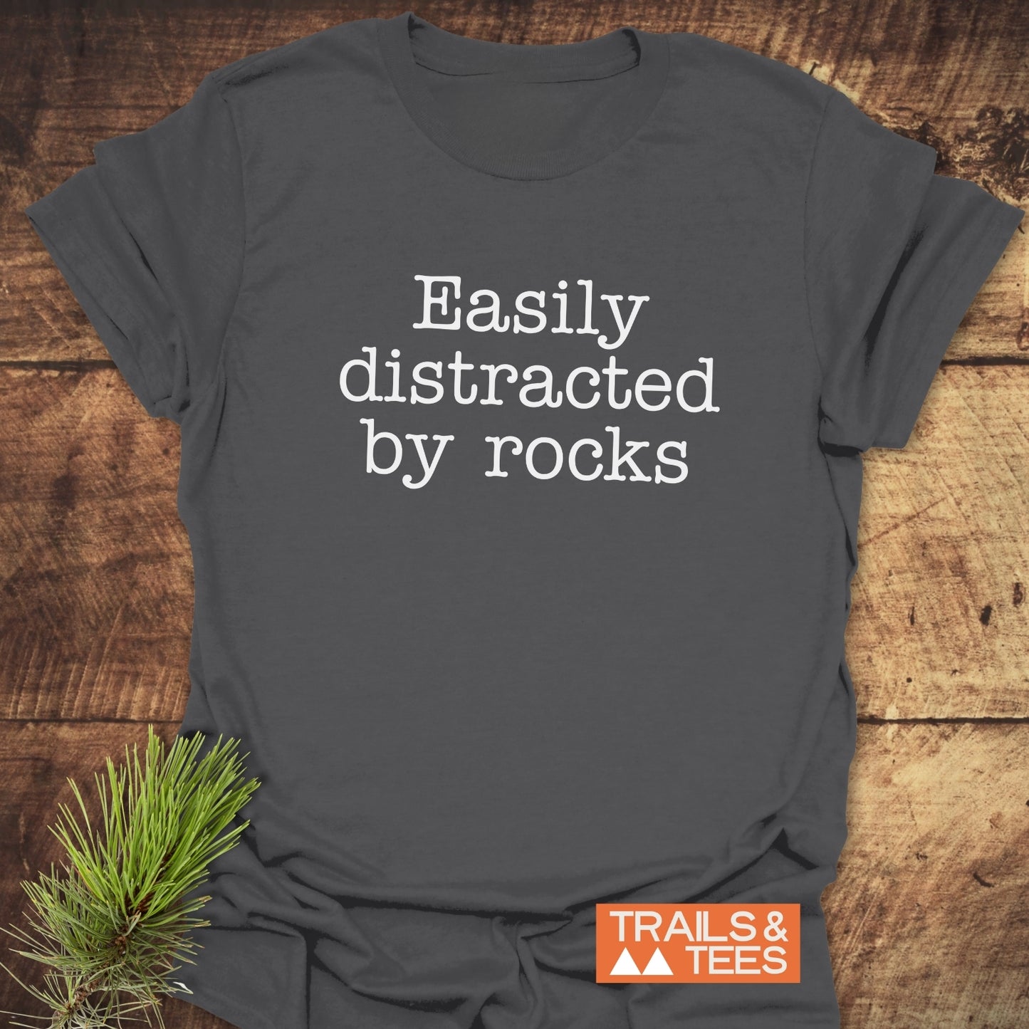 Easily Distracted Rocklover T-Shirt on wood, featuring white typewriter text. Includes TRAILS & TEES logo and pine bough, highlighting outdoor adventure spirit.