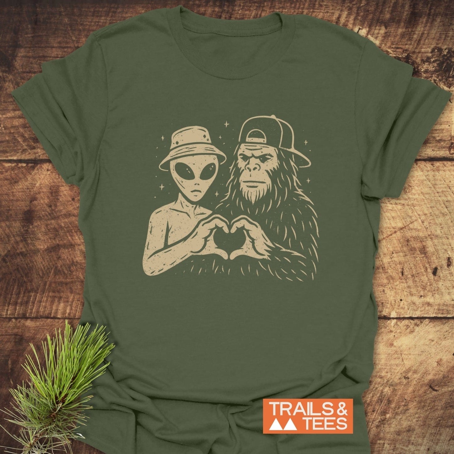 Strange Love Bigfoot T-Shirt featuring an alien and Bigfoot forming a heart shape, logo visible. Soft, durable cotton for outdoor adventures.