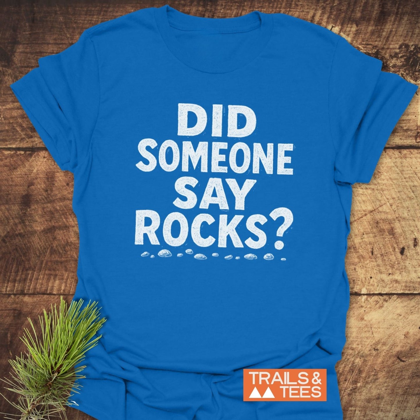 Bright blue Did Someone Say Rocks? T-shirt with pebble illustrations and TRAILS & TEES logo, laid on wood with pine sprig.