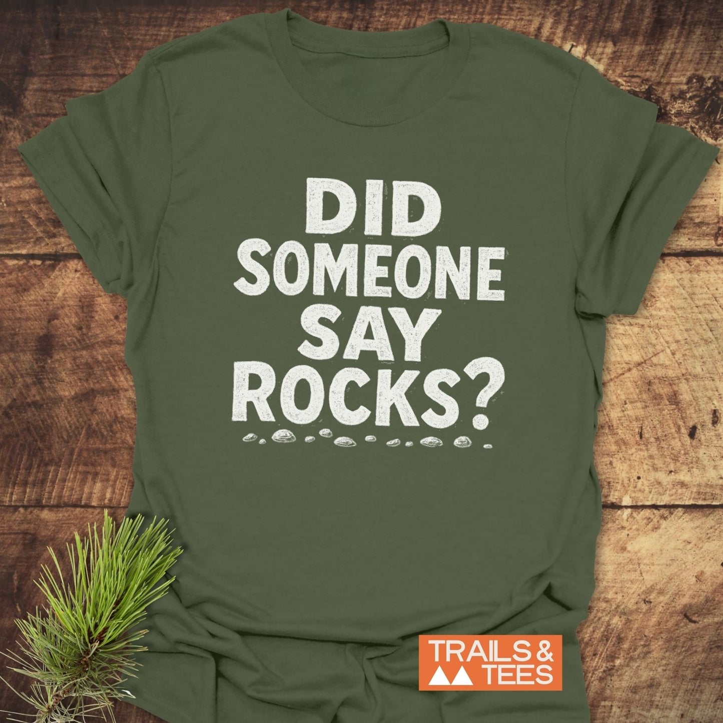 Did Someone Say Rocks? T-Shirt on wood, showcasing bold white text and rock sketches. Includes pine sprig, emphasizing outdoor theme and comfort-focused design.