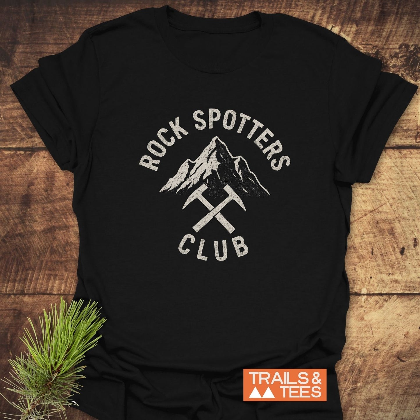 Rock Spotters Club T-Shirt featuring a vintage mountain graphic with crossed rock-hammers, laid on wood surface with pine sprig; crafted from lightweight cotton.