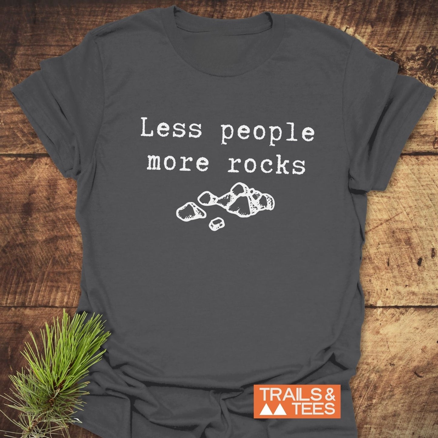 Dark-gray Less People More Rocks T-shirt with typewriter-style text and rock drawing, featuring a TRAILS & TEES label, laid on wooden surface.