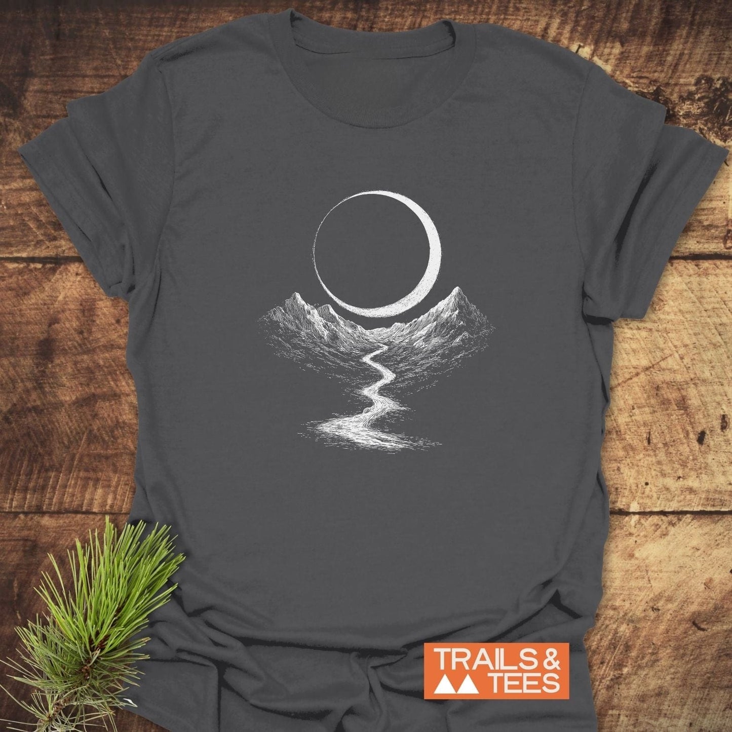 Eclipse Trail T-Shirt with a crescent moon and mountain design, laid on wood, showcasing outdoor-inspired comfort and durability with DTF prints.