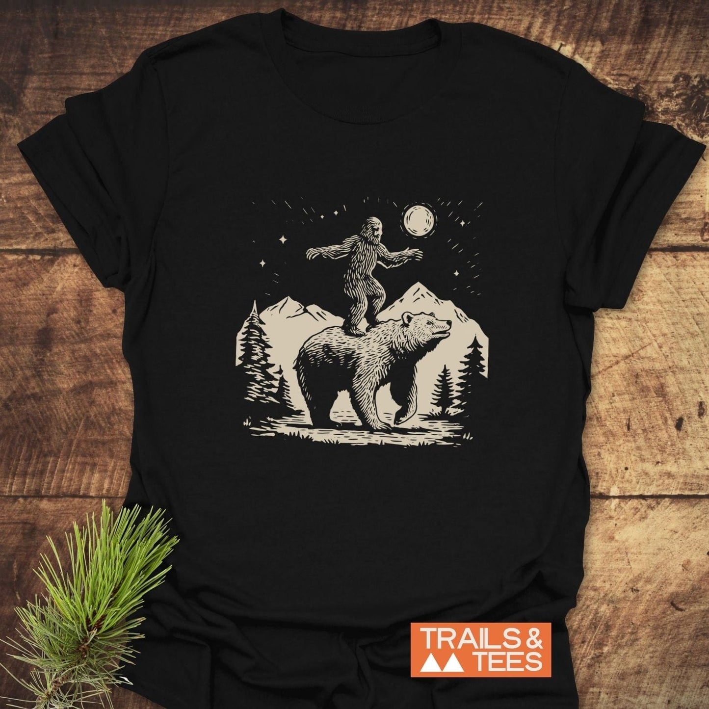 Bigfoot And Bear Funny T-Shirt featuring a whimsical forest scene design on a black tee, displayed with a pine branch, highlighting outdoor adventure themes.