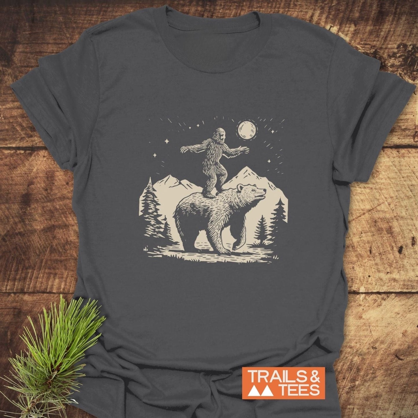 Bigfoot And Bear Funny T-Shirt on wood surface, showcasing Sasquatch balancing on a bear, with mountain scenery. Features soft, durable cotton for outdoor enthusiasts.