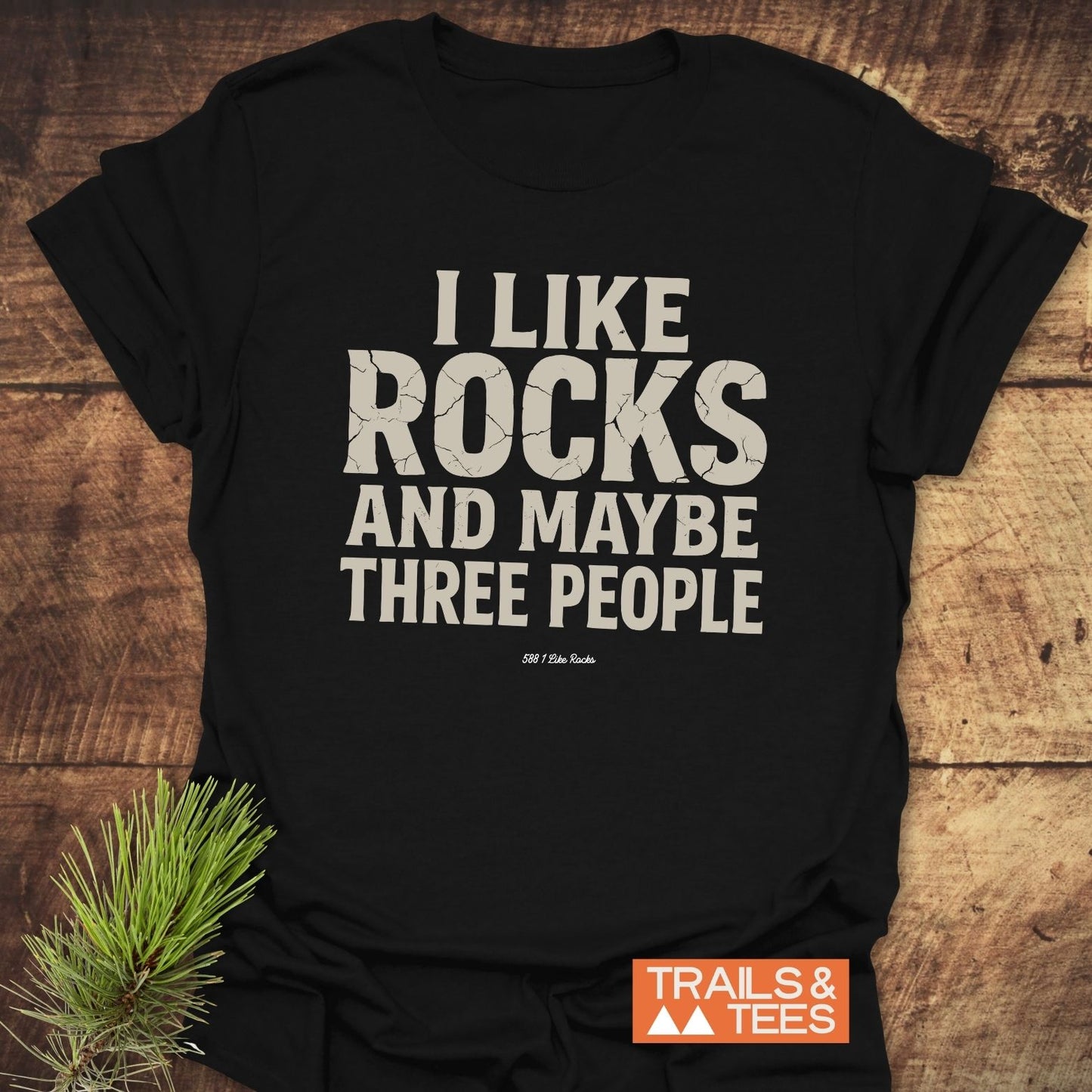 I Like Rocks T-Shirt featuring cracked-style lettering on soft cotton, laid on wood with pine sprig; Trails & Tees logo visible, perfect for outdoor enthusiasts.
