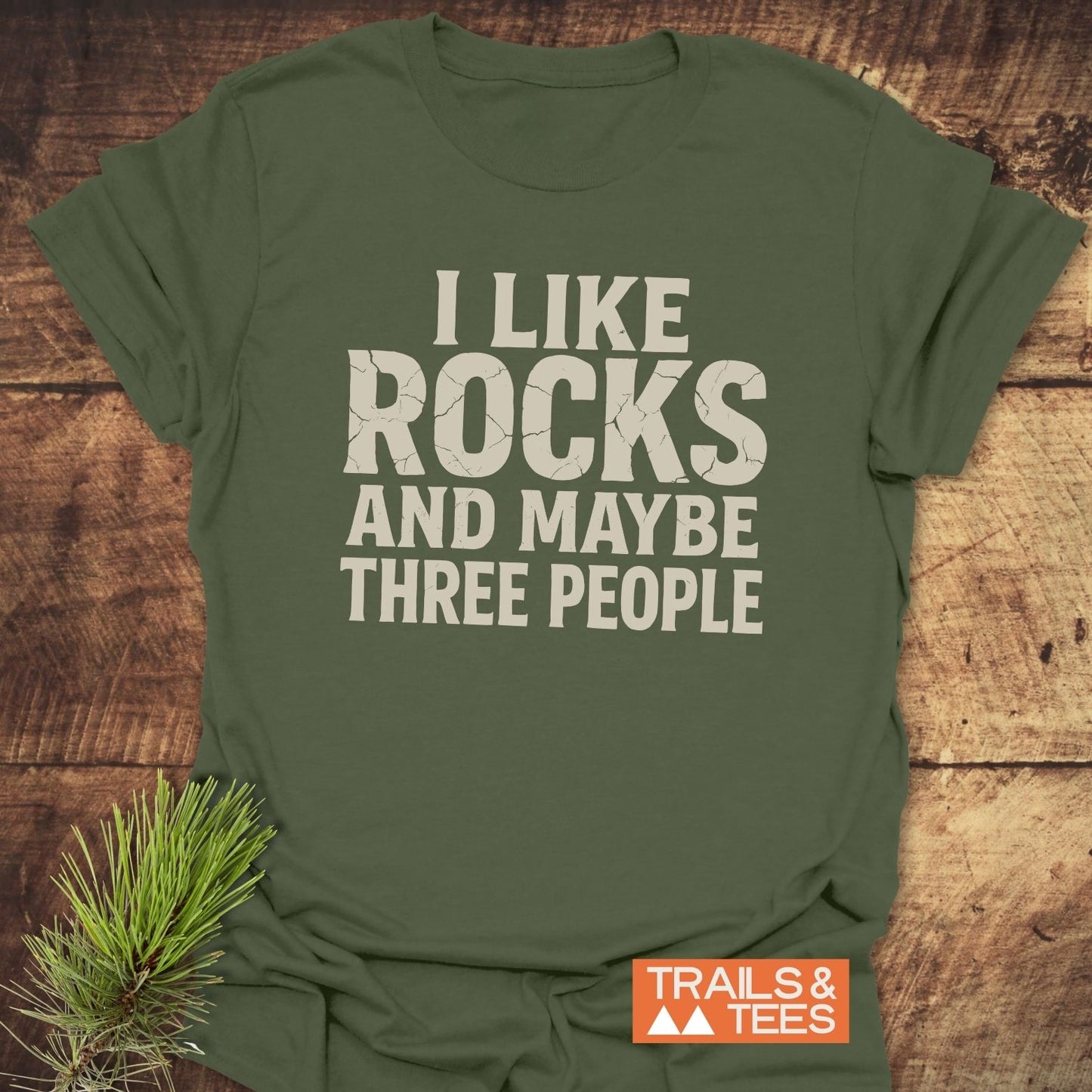I Like Rocks T-Shirt with I LIKE ROCKS AND MAYBE THREE PEOPLE print, featuring Trails & Tees logo, displayed on wood with pine sprig.