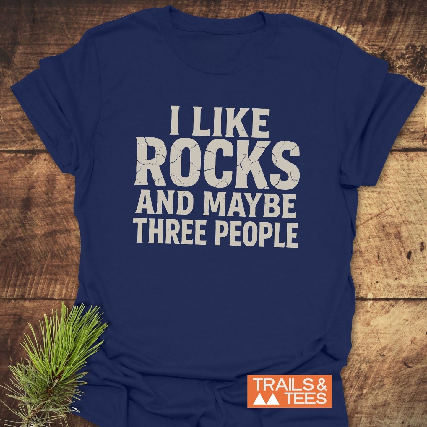 Navy I Like Rocks T-shirt with white cracked lettering, featuring a Trails & Tees logo, laid on wood with pine sprig, highlighting outdoor-inspired design.