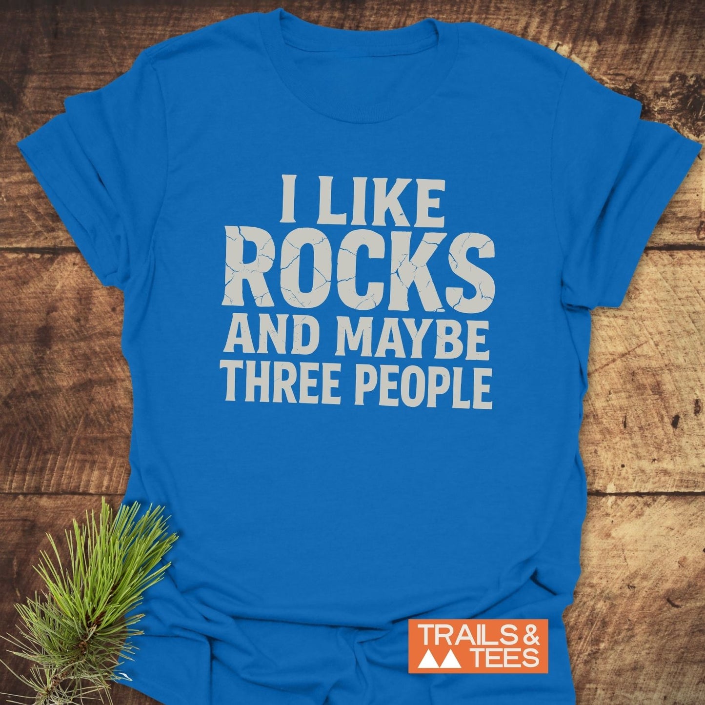 Bright blue I Like Rocks T-Shirt with white text, Trails & Tees logo, pine sprig accent, showcasing comfortable, durable, ring-spun cotton for outdoor adventures.