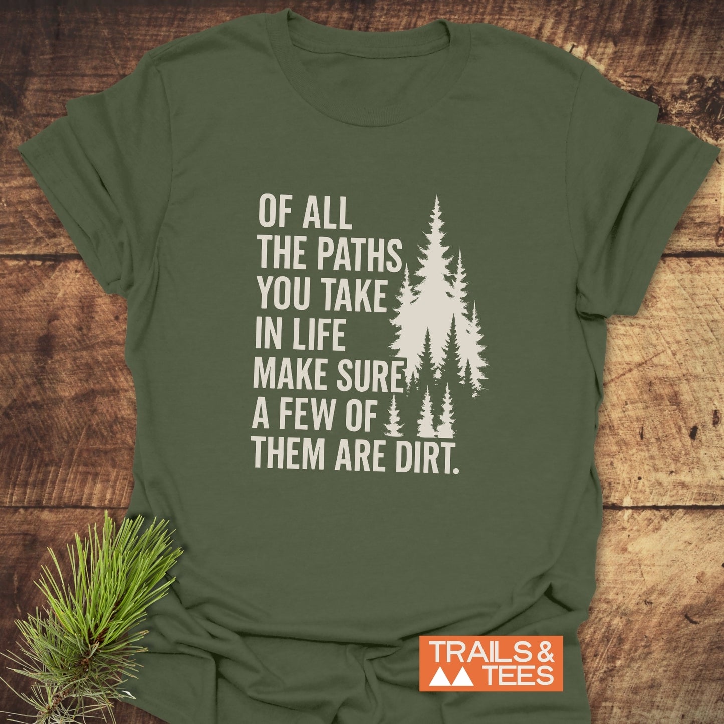 Olive-green Of All The Paths T-Shirt with outdoorsy slogan and pine graphics, styled on wood with a pine sprig accent, highlighting trail-inspired adventure.
