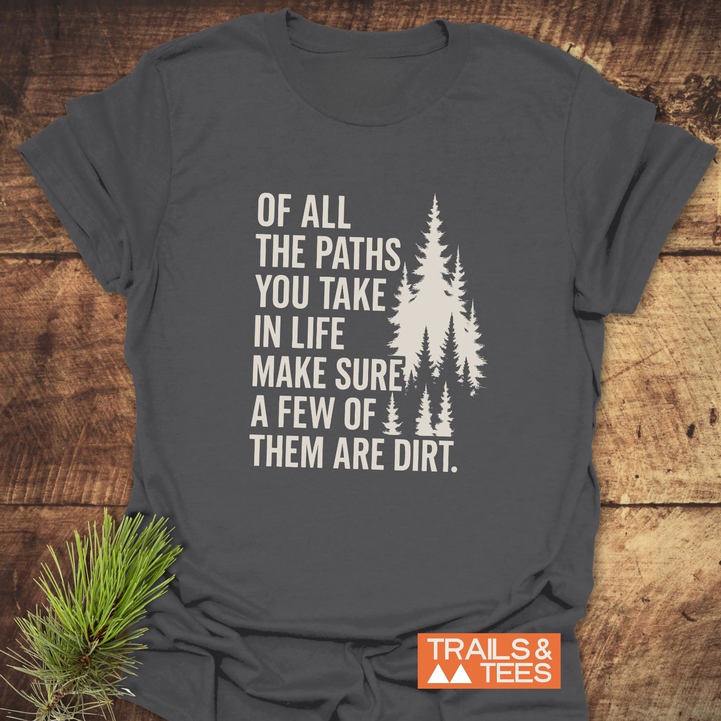 Dark-gray Of All The Paths T-Shirt with evergreen tree print and TRAILS & TEES. logo, highlighting adventure-inspired design and comfort on a wooden surface.