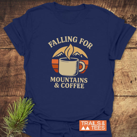 Falling For T-Shirt with coffee mug and mountain graphic, slogan “FALLING FOR MOUNTAINS & COFFEE.” Laid on wood surface, Trails & Tees logo visible.