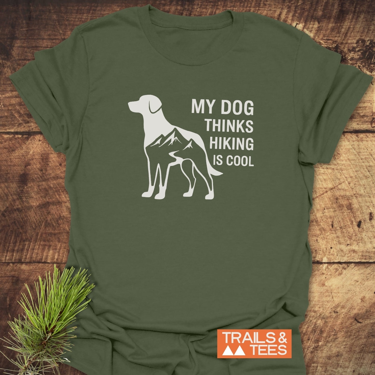 Dog Thinks... T-Shirt on wood surface, featuring a dog silhouette with mountain design and slogan. Soft, durable cotton, pre-shrunk, no side seams, brand: TRAILS & TEES.