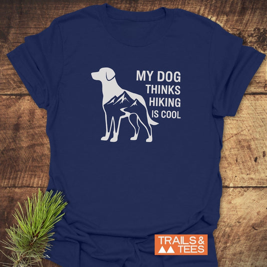 Dog Thinks... T-Shirt featuring a dog silhouette with mountain trail design, MY DOG THINKS HIKING IS COOL, on soft, pre-shrunk cotton beside a pine sprig.