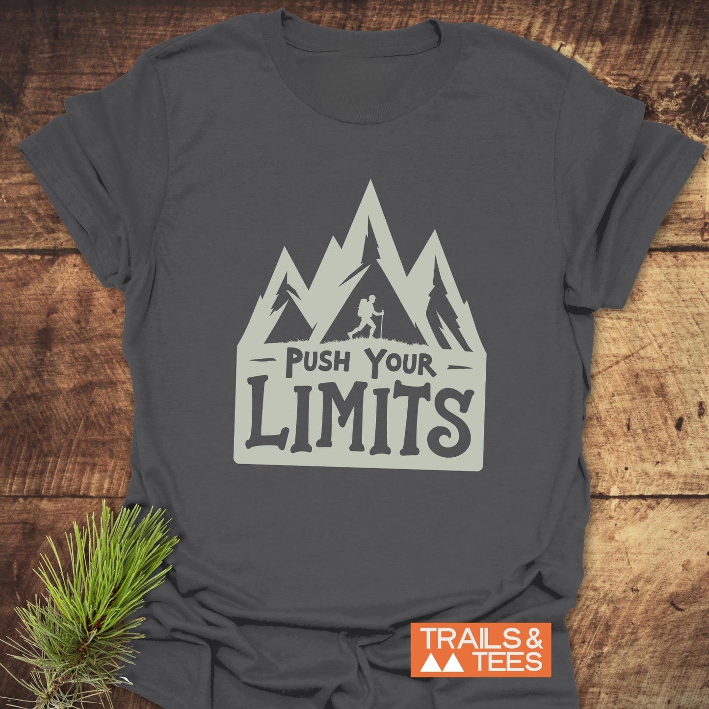 Push Your Limits T-Shirt with mountain silhouette and backpacker print, laid on wood surface, featuring TRAILS & TEES logo and pine needles for outdoor touch.