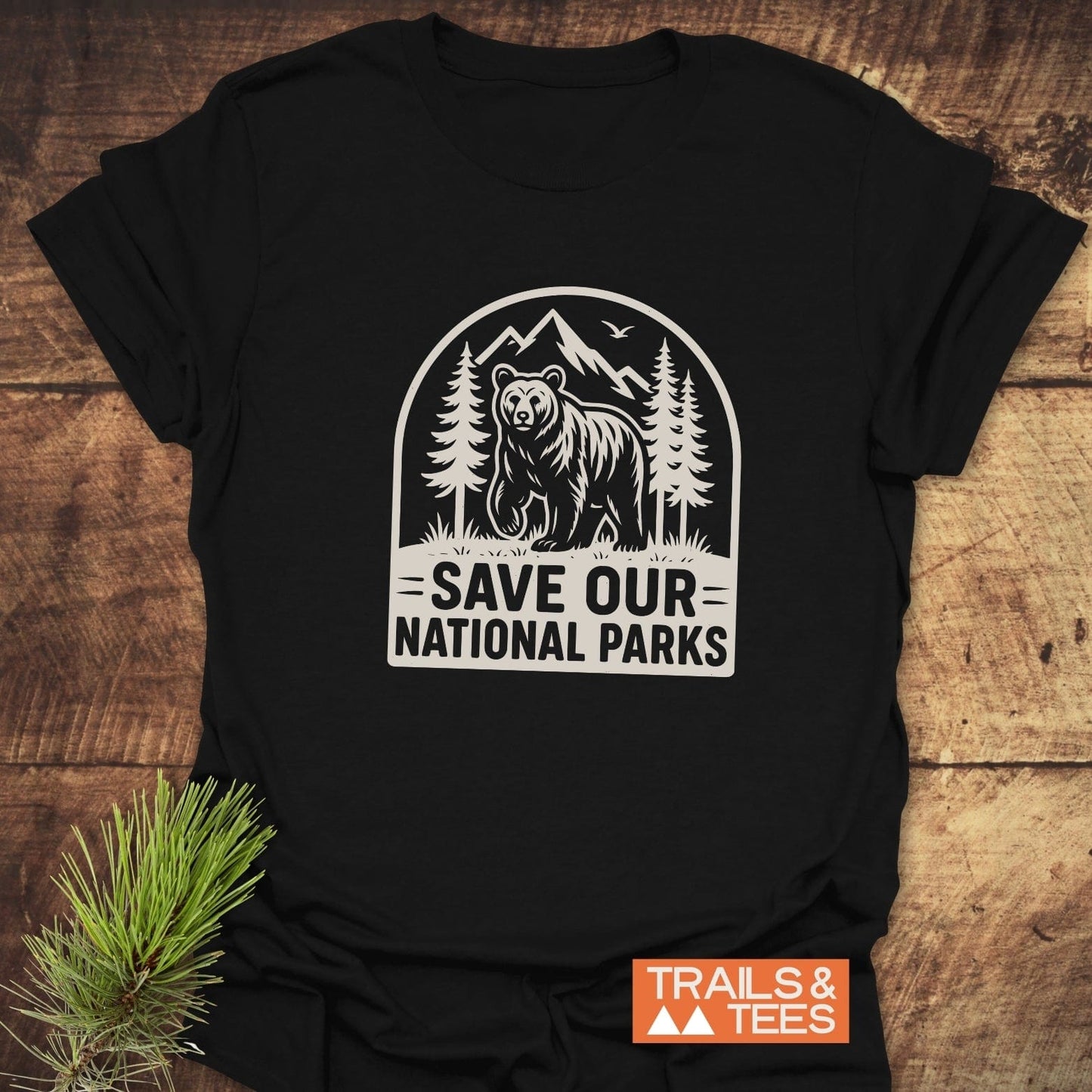 Save National Parks T-Shirt featuring a bear among trees and mountains, with a slogan and TRAILS & TEES logo, laid on a wooden surface.