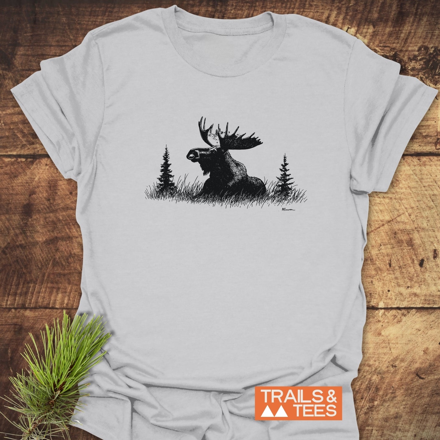 Relaxing Moose T-Shirt with a moose and pine trees design on light-gray fabric, featuring the TRAILS & TEES logo, styled with a pine branch.