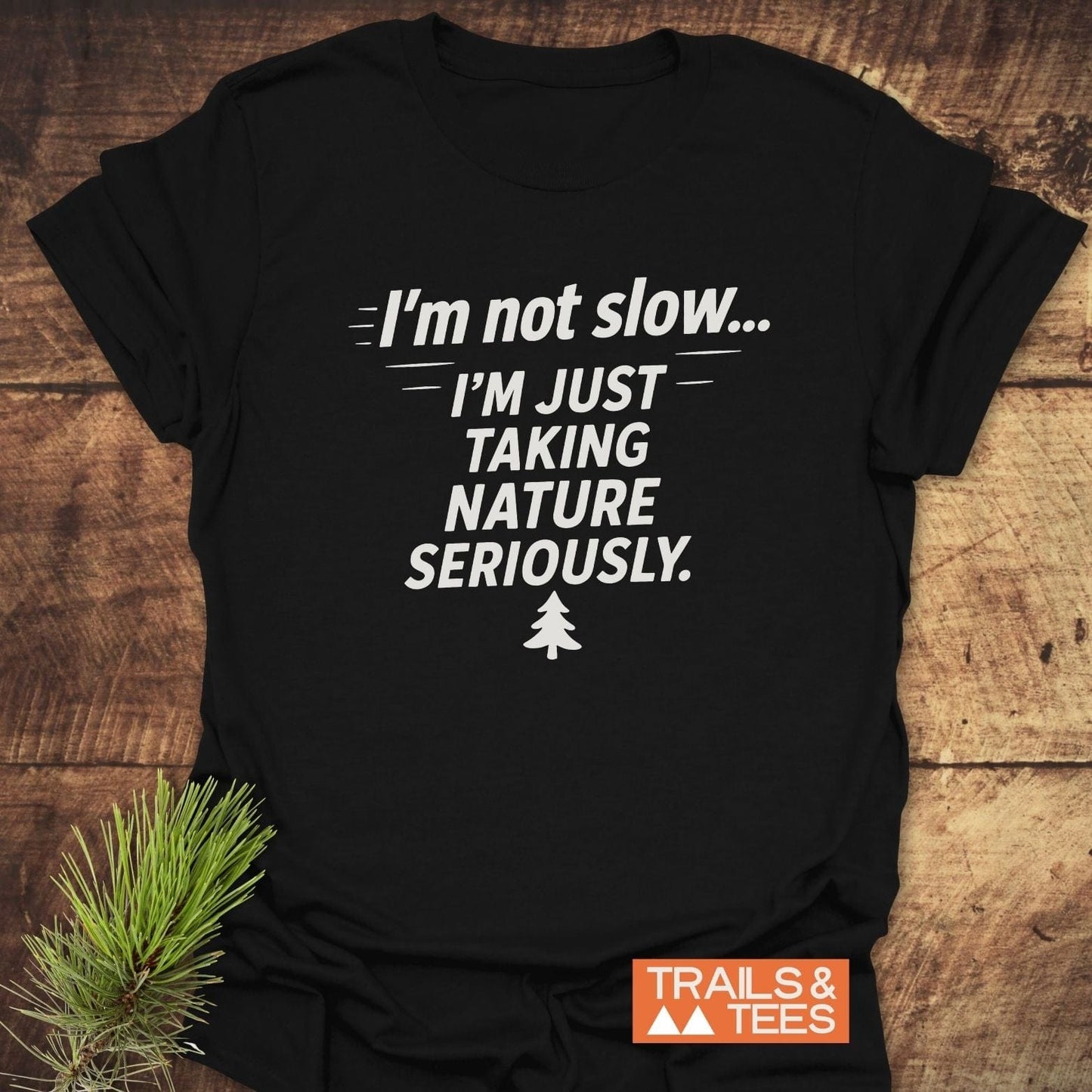 I'm Not Slow... Funny T-Shirt featuring nature-themed slogan and pine-tree icon, laid on a wooden surface with pine sprig, showcasing outdoor-inspired design.