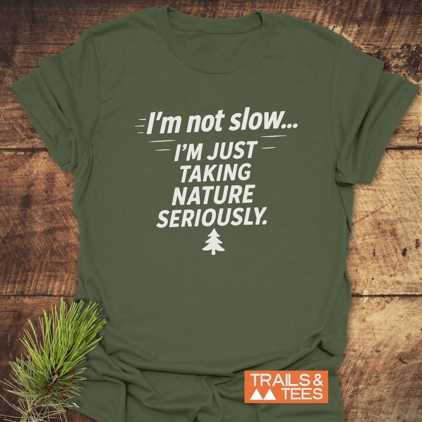 Olive-green I'm Not Slow... Funny T-Shirt with nature-themed slogan and pine-tree icon, displayed with pine sprig accent on rustic surface.