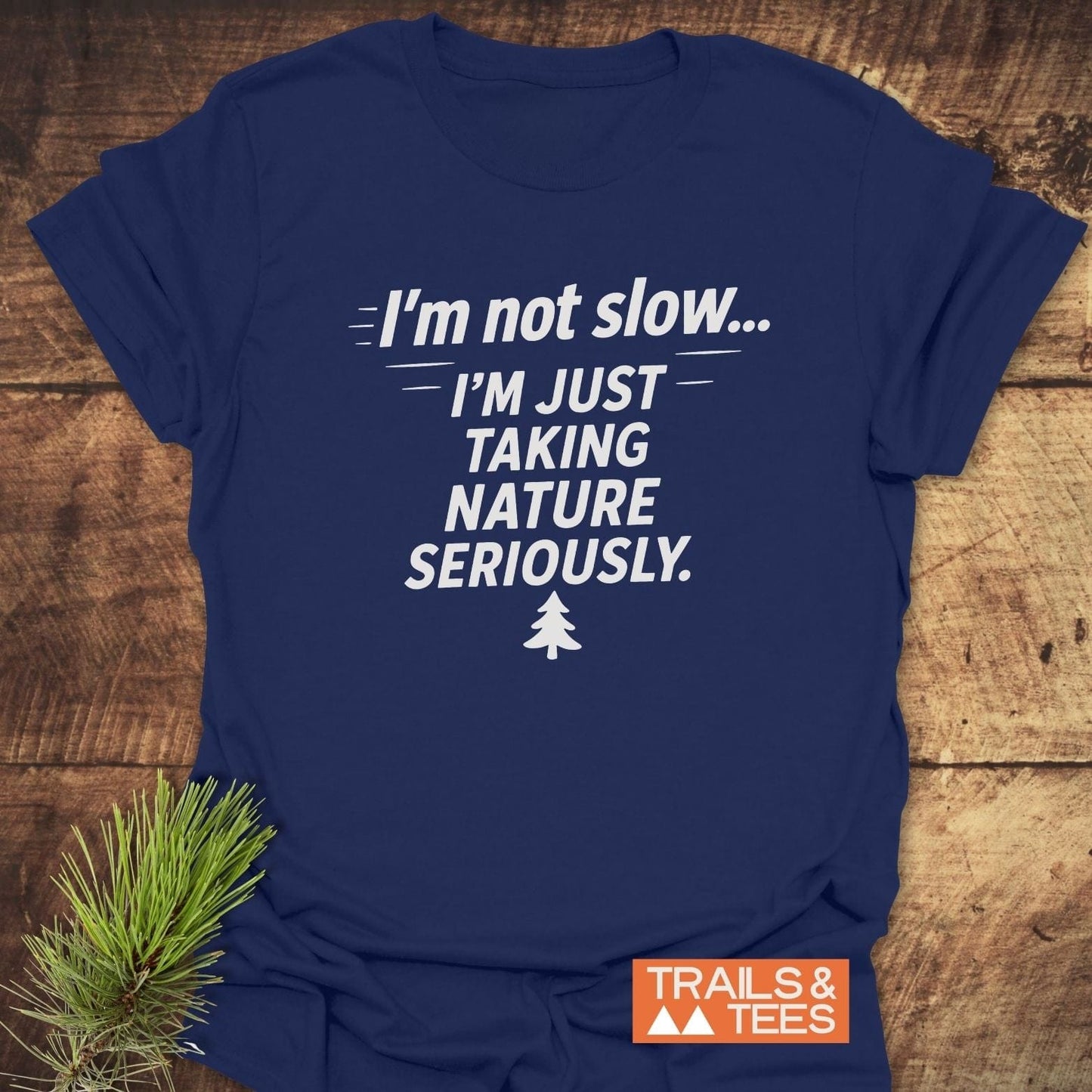 Navy T-shirt with “I’m Not Slow… I’m just taking nature seriously” slogan and pine-tree icon, by TRAILS & TEES, on wooden surface with pine branch.
