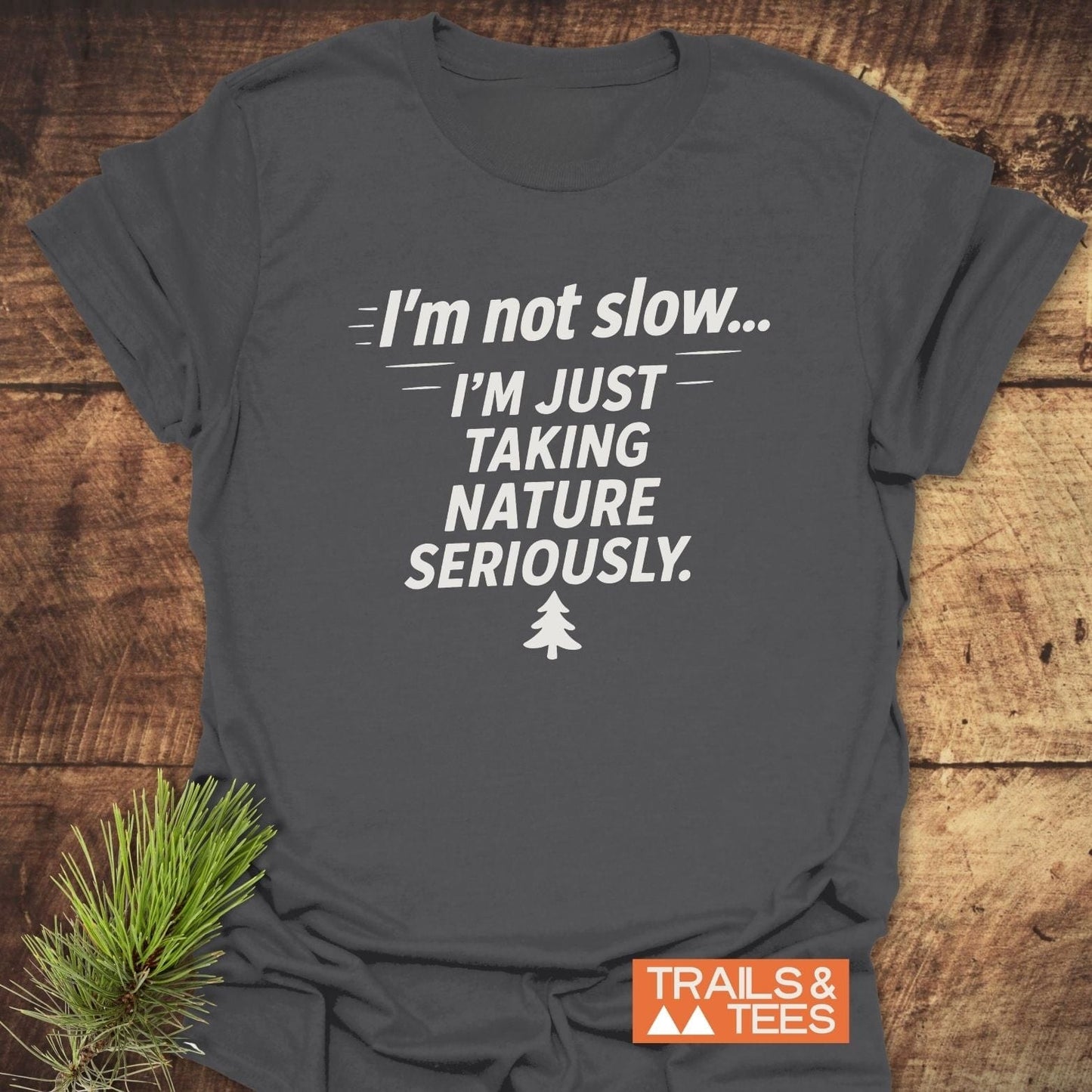 Charcoal-gray I'm Not Slow... Funny T-Shirt with white slogan and pine icon, laid flat with a pine sprig, showcasing Trails & Tees logo.